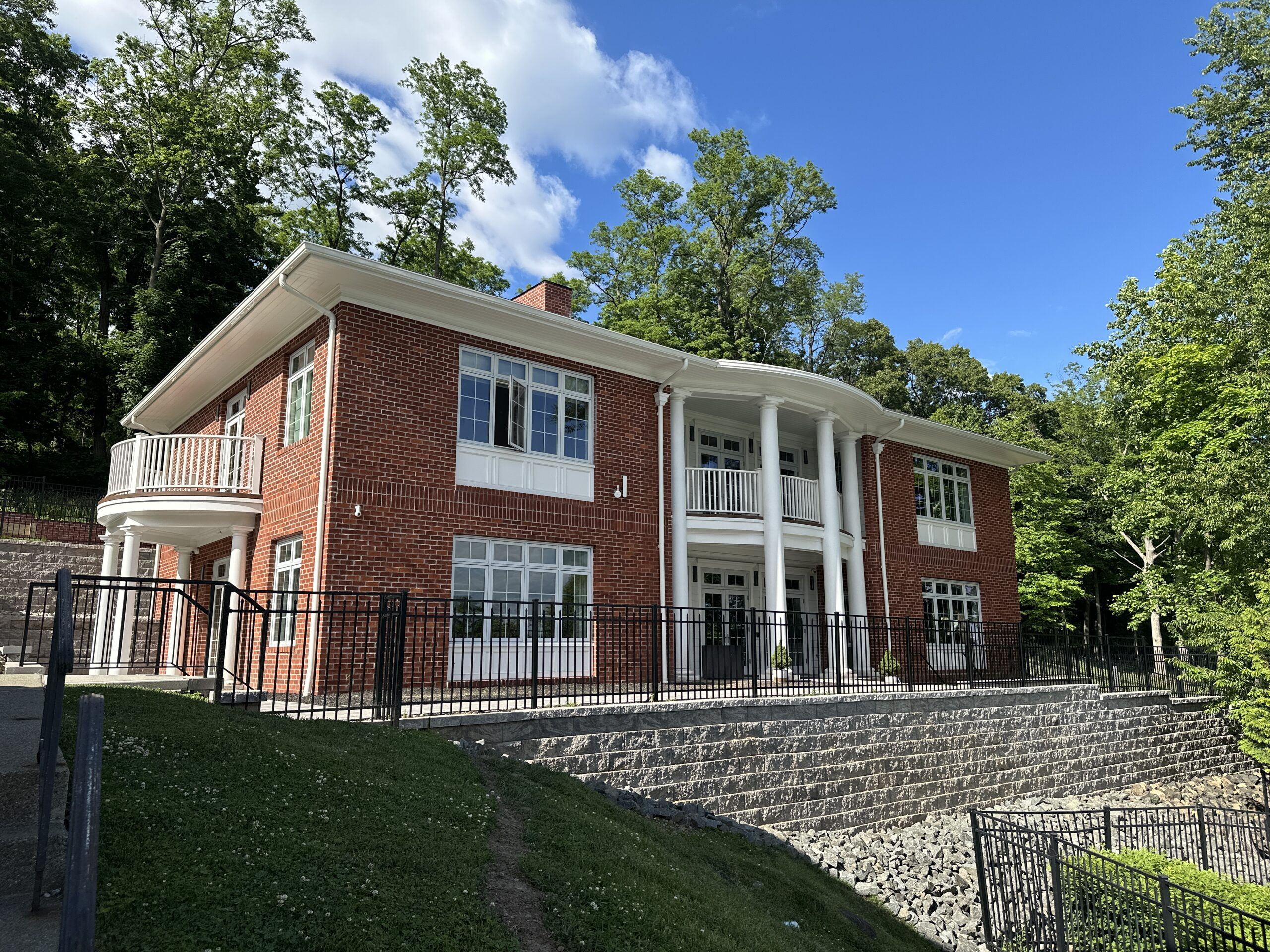 Brick two-story building with white columns, curved balcony, and large windows under a blue sky with surrounding trees.