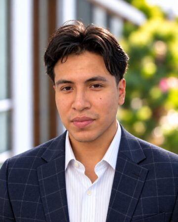 Man in a navy check blazer and white striped shirt posing outdoors with greenery in the background.