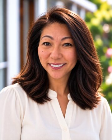 Smiling woman with dark brown hair in a white blouse, outdoors with blurred greenery in the background.