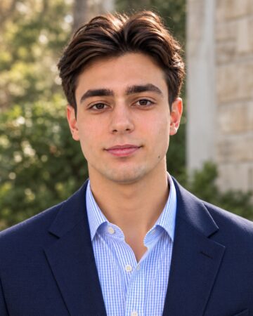 Head-and-shoulders portrait of a young man in a navy blazer and light blue striped shirt, posing outdoors with a neutral expression.