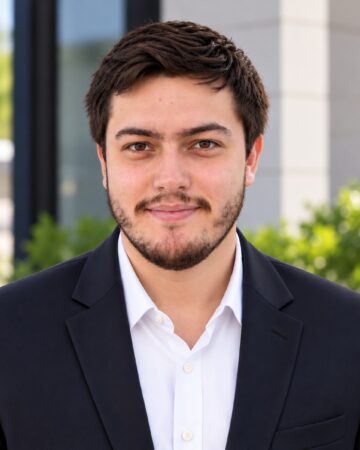 Portrait of a young man with dark hair and light beard wearing a navy blazer and white shirt, outdoors.